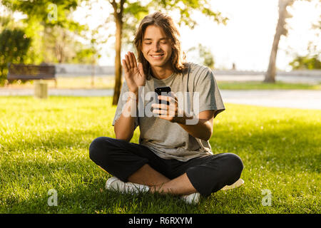 Immagine del giovane ragazzo 16-18 in abbigliamento casual seduto sull'erba verde parka nd utilizzando il telefono cellulare per le chat video Foto Stock