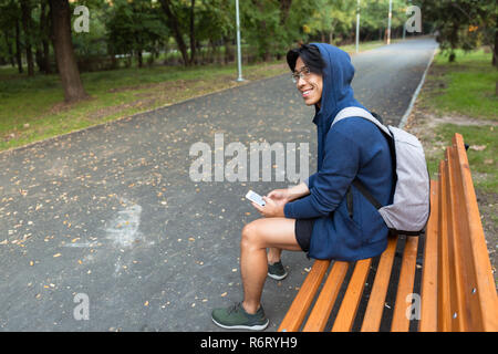 Sorridente giovane uomo asiatico vestito in felpa con cappuccio seduta su una panchina nel parco, portando allo zaino, utilizzando il telefono cellulare Foto Stock