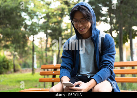 Allegro giovane uomo asiatico vestito in felpa con cappuccio seduta su una panchina nel parco, portando allo zaino, utilizzando il telefono cellulare Foto Stock