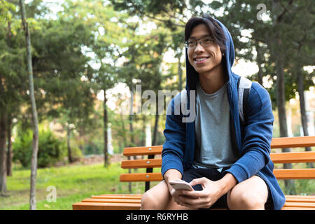 Felice giovane uomo asiatico vestito in felpa con cappuccio seduta su una panchina nel parco, portando allo zaino, utilizzando il telefono cellulare Foto Stock