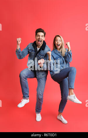 A piena lunghezza Ritratto di una felice coppia giovane vestito in abiti denim isolate su sfondo rosso, celebrando il successo Foto Stock