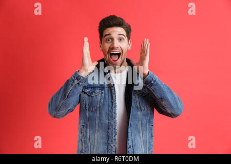 Ritratto di un allegro giovane uomo vestito con una camicia di denim in piedi isolato su sfondo rosso, urlando Foto Stock