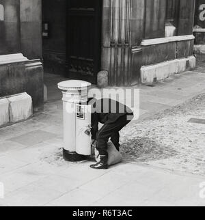 Degli anni Cinquanta, storico, un postino con il suo sacco piegarsi per svuotare una casella postale, Londra, Inghilterra, Regno Unito. Foto Stock