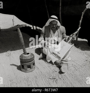 Degli anni Cinquanta, storico, un arabo beduino indossando un keffiyeh headress seduti su un tappeto di reed all'interno della sua tenda a giocare un rebab, un beduino tradizionale strumento musicale, noto come il deserto violino. Il rebab è costituito da un rettangolo di asse di legno ricoperta di pelle - una scatola sonora- e attraverso questo una singola stringa da un cavallo e di coda un crine prua per riprodurlo. Tradizionalmente utilizzato per accompagnare letture di poesie e fortemente connesso con la tribù beduina, la storia e i suoi antenati, il rebab è di grande importanza e molto apprezzato anche nella loro cultura. Foto Stock