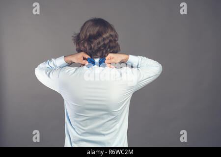 Il sig. perfezione. Vista posteriore del bel giovane uomo che indossa maglietta blu regolando il suo bow tie rimanendo contro uno sfondo grigio. Elegantemente vestito guy. Tonica. Copia dello spazio. Foto Stock