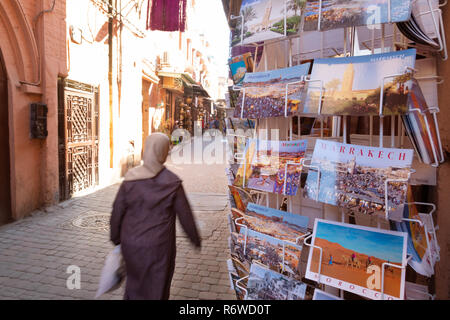 Marrakech turismo - una donna locale cammina davanti a un negozio di vendita di cartoline nel souk, Medina di Marrakesh, Marocco Africa del Nord Foto Stock