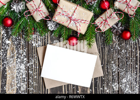 Bianco Natale greeting card e la busta con abete rami, confezioni regalo, decorazioni e pigne di neve sul telaio in legno scuro dello sfondo. moc Foto Stock