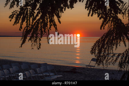 Sedie a sdraio sulla spiaggia al tramonto sul lago Ontario dopo un matrimonio. I rami degli alberi profilarsi come set di sole con colori drammatici sull orizzonte. Foto Stock