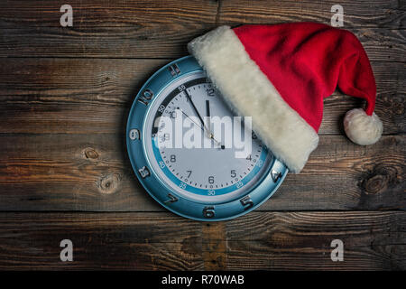 Concetto: Natale e il nuovo anno. Santa è il cappello indossato sull'orologio a muro e le frecce mostrano ormai imminente nuovo anno di mezzanotte su sfondo di legno con copia s Foto Stock