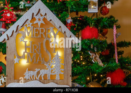 Presepe in legno design con buon Natale messaggio prima di albero di lit. Un legno intagliato presepe illuminato con una scena invernale contro l albero di Natale artificiale. Foto Stock