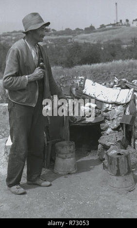 1930s, storico, Cecoslovacchia, sudentenland, pre-ww2 e un disheveled l uomo con il suo longpipe bavarese in piedi al di fuori di un basso livello di baracca in terreno accidentato, più eventualmente un senzatetto peson, un girovago o barbone. Foto Stock