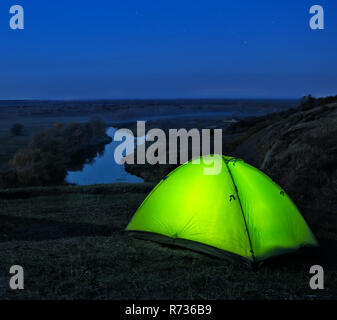 Illuminati dall'interno tenda verde su una collina sopra il fiume. Paesaggio notturno. Il concetto di libertà, privacy e viaggi. Foto Stock