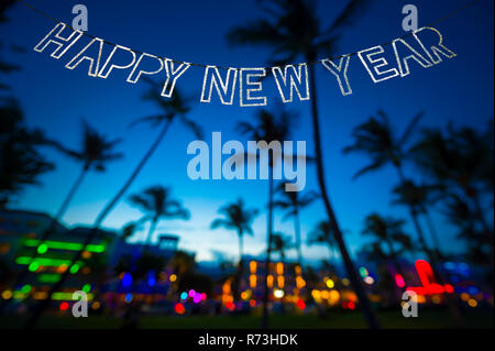 Felice Anno Nuovo messaggio appesi in glittery parte bunting al di sopra di alberi di palma tropicali dell'incandescente luci al neon della skyline di Miami Beach, Florida Foto Stock