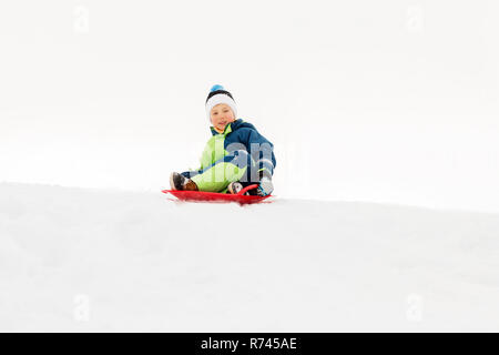 Felice ragazzo scorrevole sulla slitta verso il basso la collina di neve in inverno Foto Stock