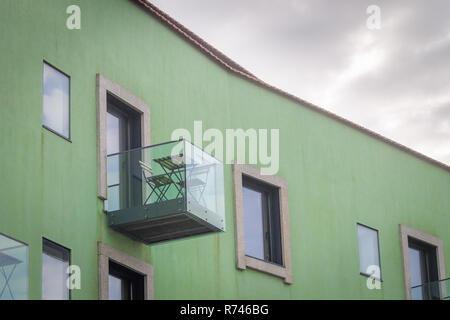 Vista laterale di un bicchiere di balcone con tavolo e sedie in legno, su un dipinto di verde edificio. Giorno nuvoloso. Foto Stock