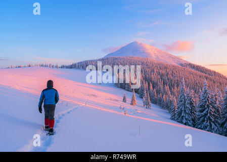 Il ragazzo nel down jacket passeggiate lungo il sentiero innevato. Paesaggio invernale con una bella montagna. Tramonto colorato Foto Stock