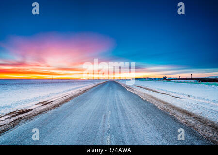 Asfalto smerigliato paese strada attraverso inverno campi innevati e prati al tramonto o l'alba. Paesaggio in Bielorussia o parte europea della Russia Foto Stock