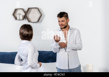 Bel ragazzo abbottonatura bracciale e guardando la fidanzata abbottonatura suo shirt nella mattina dei giorni feriali in camera da letto, concetto femminista Foto Stock