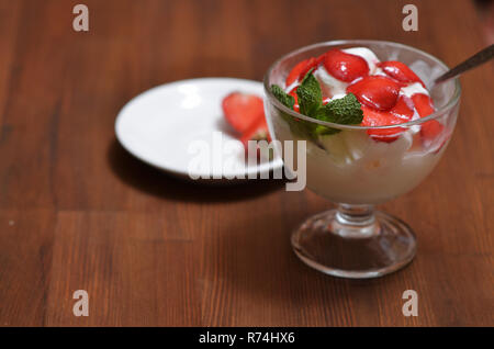 Gelato alla vaniglia con fragole a fette e foglie di menta in una ciotola di vetro con un cucchiaio su una tavola di legno Foto Stock