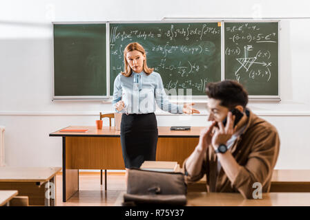 Insoddisfatto maestro guardando uno studente che parla sullo smartphone in aula Foto Stock