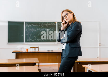 Bella pensieroso insegnante femminile in abbigliamento formale e bicchieri in aula Foto Stock