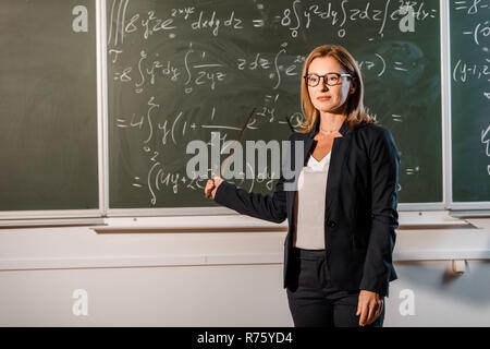 Bellissima femmina insegnante con puntatore in legno spiegando equazioni matematiche in aula Foto Stock