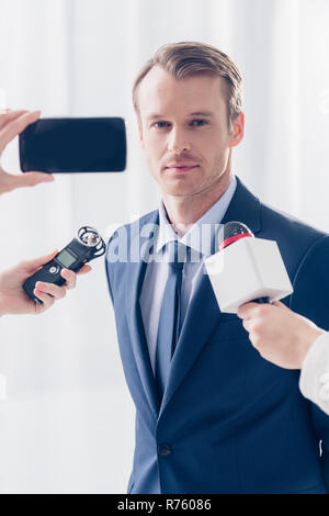 Bello imprenditore dando intervista a giornalisti e guardando la telecamera in ufficio Foto Stock