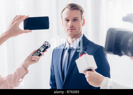 Bello imprenditore dando intervista a giornalisti e guardando la telecamera in ufficio Foto Stock