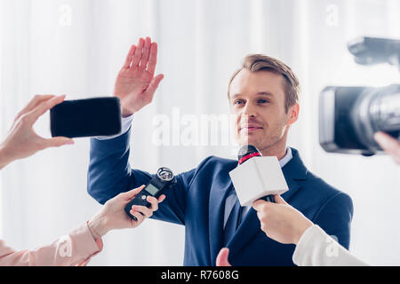 Bello imprenditore dando intervista ai giornalisti in ufficio e agitando la mano Foto Stock