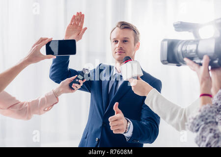 Bello imprenditore dando intervista ai giornalisti in ufficio, agitando la mano e mostrando il pollice in alto Foto Stock