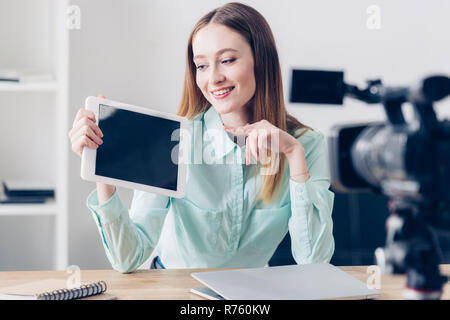 Sorridente femmina attraente video registrazione blogger vlog e puntando su tablet con schermo vuoto in ufficio Foto Stock