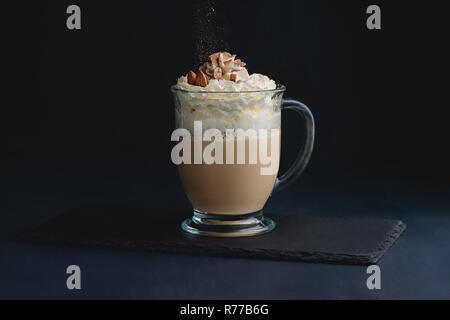 Il caffè e il latte da bere latte macchiato con panna montata, dadi e cacao in polvere. Dolce bevanda calda su sfondo scuro con copia spazio per un menu Foto Stock