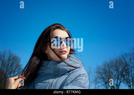 Uno stile di vita all'aperto close up ritratto della bella ragazza camminare in inverno nevoso park. Sorridente e godendo d'inverno. Indossare Eleganti occhiali da sole con mirroring, blu trench coat Foto Stock