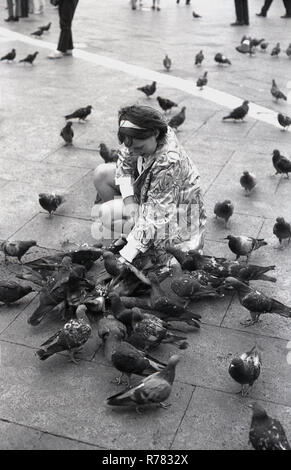 1970, una giovane ragazza inginocchiata circondata da piccioni in Piazza Sam Marco o Piazza San Marco, Venezia, Italia. I piccioni sono sempre stati una popolare attrazione turistica a Venezia e una volta hanno rivaleggiato i gatti come le mascotte tradizionali, se non ufficiali, della città lagunare. Foto Stock