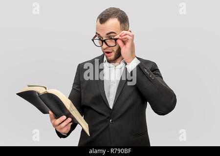 Si chiedeva ed entusiasta giovane tenere libro in una mano e i bicchieri con un'altra. Egli guarda nel libro. Ragazzo indossa tuta. Isolared su sfondo bianco. Foto Stock