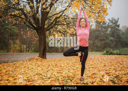 Calma, pacifica e positiva giovane donna sorge in posizione asana e guardare dritto. Essa tiene in alto le mani. La donna è in autunno park da solo. Foto Stock