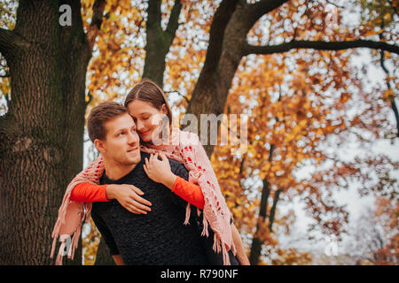 Allegro e positivo giovane donna sedersi sul ragazzo torna e si appoggia a lui. Egli mantiene il suo aspetto e per lato. Essi sono in autunno park. Foto Stock