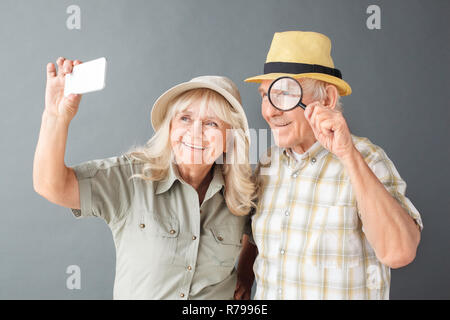 Senior turisti in spiaggia cappelli studio isolato permanente sull uomo grigio azienda magnifier giocoso tenendo selfies sullo smartphone con la moglie felice Foto Stock