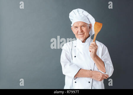 Lo chef Senior studio isolato permanente su grigio che mostra la spatola cercando positivo della fotocamera Foto Stock