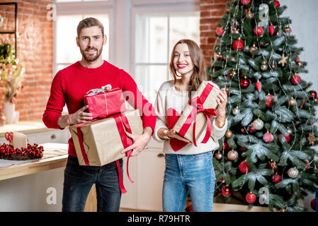Ritratto di una giovane coppia adorabile in maglioni in piedi insieme con il nuovo anno doni vicino all'albero di Natale a casa Foto Stock