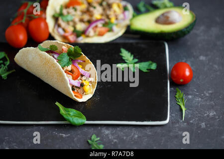 Mexican tacos con carne di maiale, spezie e verdure su una pietra nera piastra su uno sfondo scuro con ingredienti per tacos e Menu ristorante concetto. Foto Stock