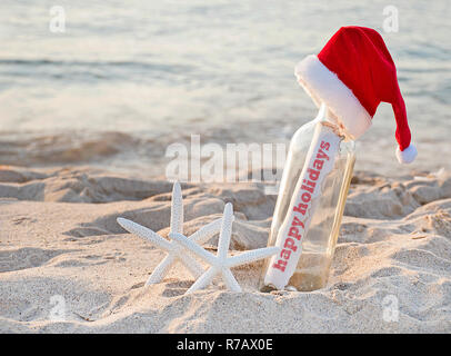 Santa hat sul messaggio in una bottiglia con un paio di bianco stella di mare nella spiaggia di sabbia e buone vacanze a tutti saluto Foto Stock