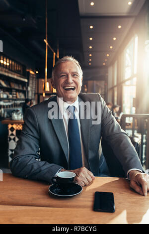Ritratto di Allegro senior uomo in tuta seduti ad un ristorante con tazza di caffè e smart phone sul tavolo. Ridendo imprenditore presso la caffetteria. Foto Stock