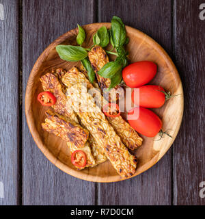 Tempeh fritto decorate con i peperoncini freschi pomodori ciliegia e le foglie di basilico sul vassoio in legno scuro su una superficie di legno. Vista dall'alto. Immagine quadrata. Foto Stock