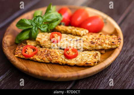 Tempeh fritto decorate con i peperoncini freschi pomodori ciliegia e le foglie di basilico su un round vassoio in legno scuro su una superficie di legno. Vista laterale Foto Stock