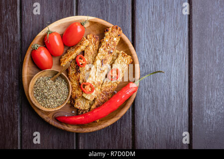 Tempeh fritto, i peperoncini freschi pomodori ciliegia e il mix di spezie asciutto in recipiente di legno con il cucchiaio di legno su una rotonda vassoio in legno Con uno spazio Foto Stock