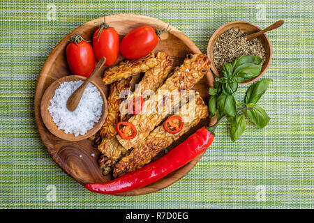 Tempeh fritto, i peperoncini freschi pomodori ciliegia, le foglie di basilico, mix di erbe aromatiche e sale marino in vasi di legno con cucchiai di legno su un round vassoio in legno Foto Stock