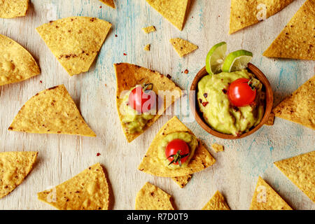 Corn chips nachos e guacamole in una ciotola di legno decorate con pomodorini e fettine di lime su un di legno bianco lavato tabella. Foto Stock