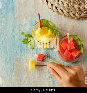 Tagliare cubetti di rosso e giallo di anguria in vasi di vetro con la menta e crostino picks su bianco e blu lavato un tavolo di legno. Cappello di paglia sullo sfondo Foto Stock