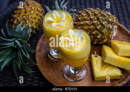 Due bicchieri di fresco succo di ananas decorata con fiori di Plumeria, due intere Ananas e albero grande pezzi tagliati su una rotonda vassoio in legno Foto Stock